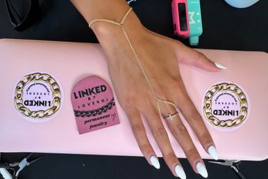 Tanned hand with white almond manicure wearing a delicate gold chain hand-harness and cross ring, resting on a pink laptop with chain-themed stickers and jewelry tools nearby