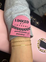 Close-up of wrist wearing two delicate gold chain bracelets with a pink 'LINKED' permanent jewelry tag resting on a gray sleeve, jewelry display in the background.