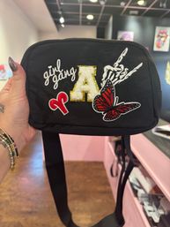 Black nylon crossbody bag held in a shop, decorated with embroidered patches reading 'girl gang', a gold-trimmed fuzzy letter A, a red butterfly and a skeleton-hand design.