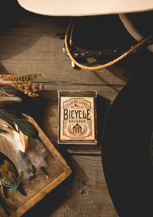 Top-down Western still life on a rustic wooden table: bourbon-themed playing card box centered, braided leather hatband and cowboy hat brim at edges, wooden tray with colorful feathers and a stone arrowhead — saloon-ready vibe.
