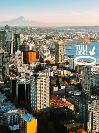 Aerial city skyline at golden hour with dense high-rise buildings, waterfront harbor, ferris wheel and stadium domes, distant snow-capped mountain; a white arrow and circled label highlight a waterfront building.