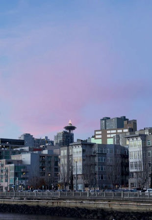 Pastel pink-blue sunset over Seattle waterfront, Space Needle peeking above modern riverside apartment buildings.
