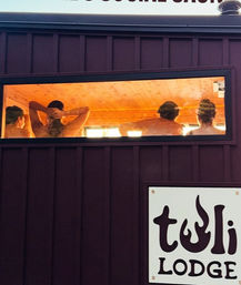 Four people relaxing in a warm, wood‑paneled sauna seen through a narrow exterior window on a dark wall, glowing orange interior with a rustic lodge spa vibe.