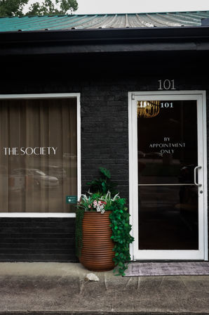 Chic black-brick storefront entrance numbered 101 with a glass door labeled “by appointment only,” adjoining window with white lettering, and a large terracotta planter spilling green foliage on the concrete sidewalk.
