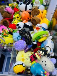 Colorful claw machine prize pile with plush toys and keychains — panda, rubber duck, burger and squishy characters inside an arcade cabinet