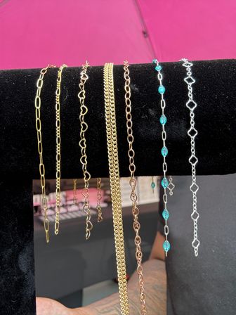 Row of gold, rose-gold and silver chain bracelets with link, heart and turquoise-beaded designs draped on a black velvet jewelry display against a bright pink backdrop.