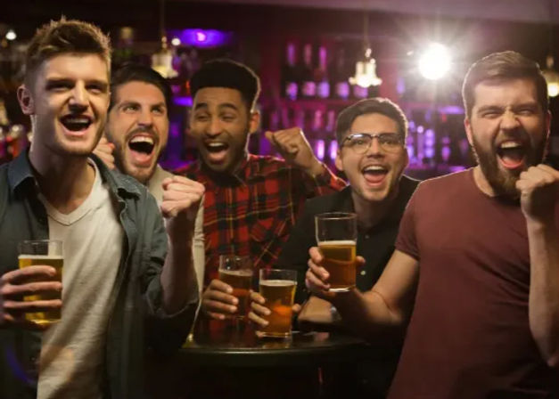 Five friends cheering with pints of beer at a lively bar — energetic night out, nightlife vibe.