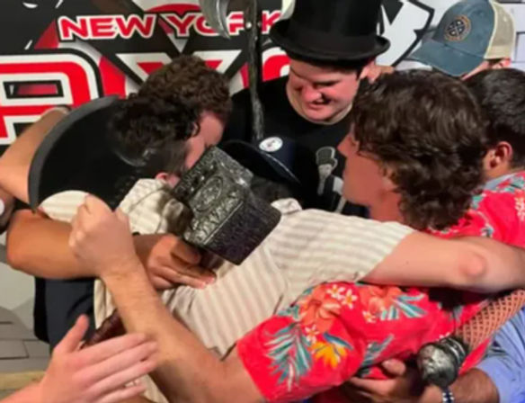 Group of friends in hats and a bright Hawaiian shirt sharing a joyful group hug on stage with an event banner in the background, New York