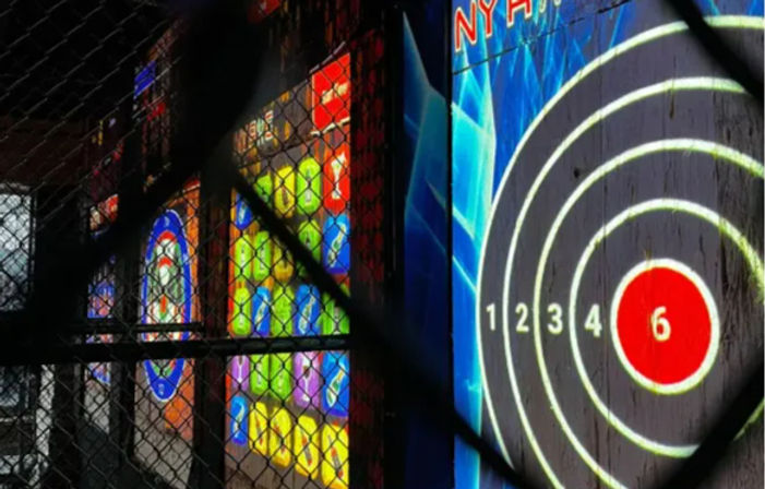 View through a chain-link safety fence of an indoor target lane with an illuminated circular scoring board showing numbered rings and a red bullseye, next to colorful arcade-style light panels.