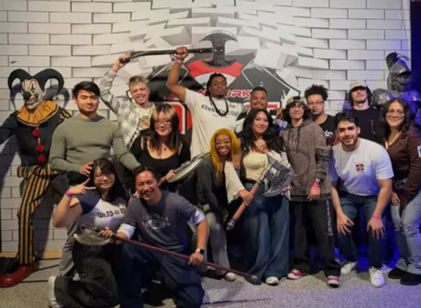 Group of friends posing at a themed indoor attraction in front of a white tiled mural, holding prop axes and a spear, flanked by a creepy clown figure and suit of armor — playful group photo from a night out.