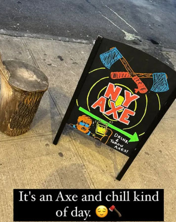 Colorful sidewalk chalkboard advertising axe‑throwing with crossed axes, concentric target and beer mug illustration, propped next to a tree stump on an urban sidewalk.