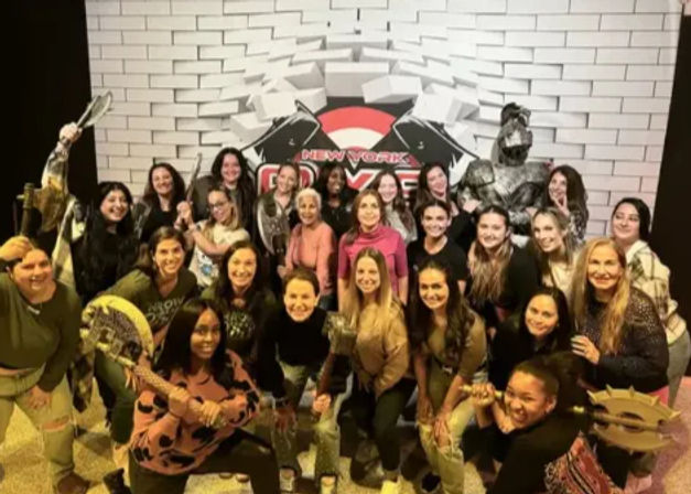Large group of smiling women playfully posing with foam axes, shields and a costumed knight in front of a white brick backdrop reading “New York” at a themed team-building event.