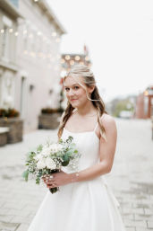 Smiling bride in a simple white gown holding a white rose and eucalyptus bouquet on a cobblestone downtown street with twinkling string lights