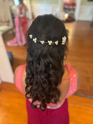 Back view of a half-up bridal hairstyle with long dark curls and a jeweled floral hair vine, indoor setting with wooden floor