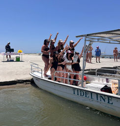 Group of women in swimsuits cheering and posing on a small party boat at a sunny sandy beach shoreline, with other beachgoers and a picnic table in the background.