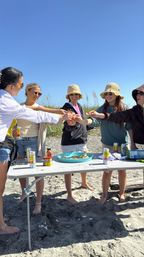 Five friends barefoot on a sunny sandy beach gathered around a folding table with seafood, drinks and condiments, smiling and toasting under a clear blue sky.