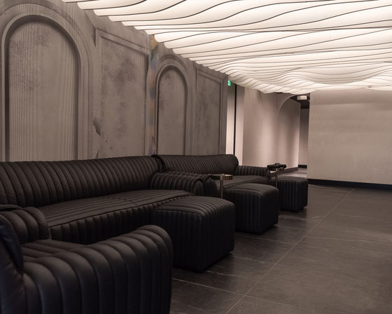 Modern minimalist lounge interior with ribbed black leather sofas and ottomans along a textured arched wall, sculptural wavy ceiling lights casting warm ambient glow over dark tile floor.