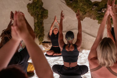 Group yoga/meditation class in a cozy wellness studio, instructor seated on a cushion with hands raised while participants mirror the pose in front of a moss-covered wall and softly glowing floor.