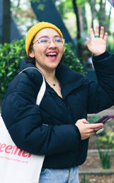 Smiling person in a yellow beanie and glasses waving while holding a smartphone and tote bag in a leafy urban park