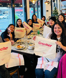 Smiling group of friends at an urban café brunch holding matching branded tote bags over a table of plates, coffee and drinks, large windows showing a busy street and a small decorated Christmas tree