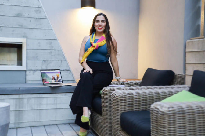 Smiling woman in business-casual outfit and bright yellow-pink scarf seated on a modern outdoor patio wicker sofa beside an open laptop under warm wall light.