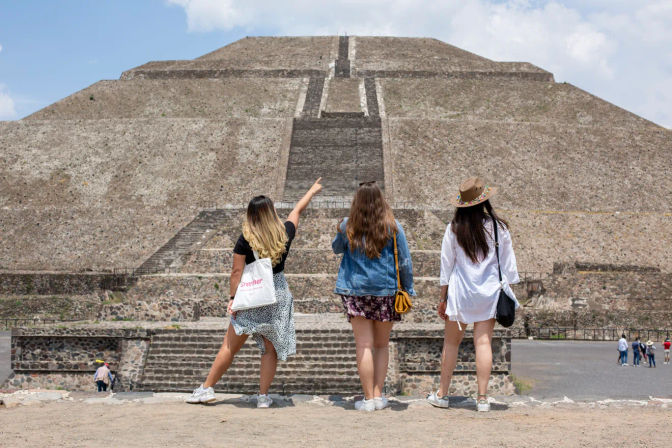 Three friends with backs to camera looking up at the massive Pyramid of the Sun at Teotihuacan, Mexico, one pointing toward the long stepped stone stairway at the archaeological site.