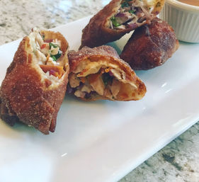 Close-up of crispy fried chicken egg rolls, halved to reveal shredded chicken, carrots and herbs, served on a white rectangular plate with a ramekin of dipping sauce on a granite countertop.