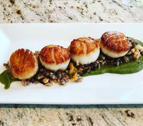 Four golden seared scallops on a bed of wild rice and vibrant green pea purée, plated in a row on a white rectangular dish over a speckled granite countertop.