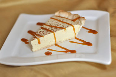 Close-up of a creamy vanilla cheesecake slice with caramel drizzle and chocolate shavings on a white square plate against a warm golden background