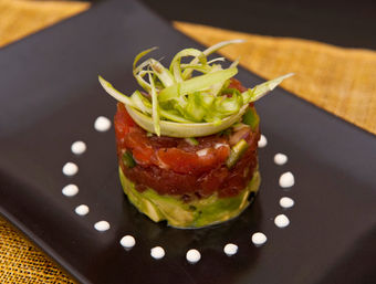 Fine-dining tuna tartare tower with avocado base, diced raw tuna and shaved green onion garnish on a black plate surrounded by dotted cream sauce
