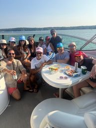 Group of friends wearing colorful caps enjoying a sunny summer pontoon boat outing on a lake, seated around a small table with snacks and drinks and a tree-lined shoreline in the background.
