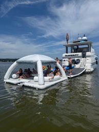 Summer boat party on a calm lake — group lounging on a white inflatable floating island tethered to a yacht under a bright blue sky with wispy clouds.