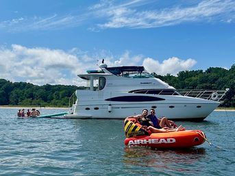 White motor yacht anchored near a tree-lined lake shoreline on a sunny summer day with people relaxing on an orange inflatable tube and a small group on the yacht's swim platform.