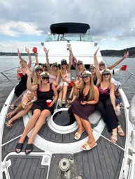 Group of women wearing captain hats and sunglasses on the bow of a white yacht on a lake, raising red cups in a summer yacht party