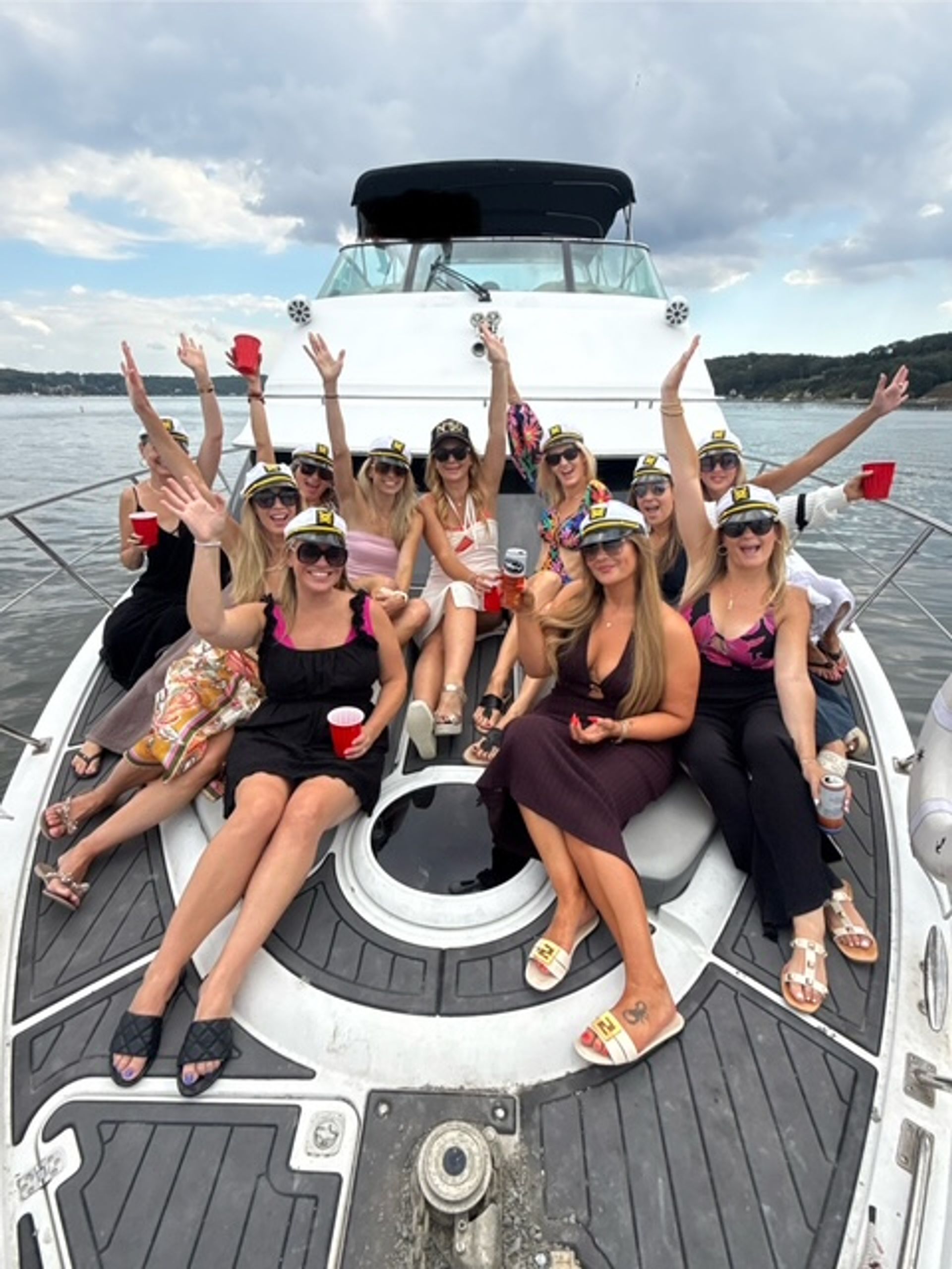 Group of women wearing captain hats and sunglasses on the bow of a white yacht on a lake, raising red cups in a summer yacht party