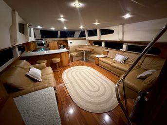 Luxury yacht interior salon with tan leather sofas and curved dinette, galley with granite countertop and bar stools, warm wood floors, oval braided rug, recessed lights and panoramic windows.