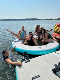 Six friends relaxing on a round inflatable float and dock at a sunny lake, wearing swimsuits, holding drinks and smiling beside a colorful towable tube on a summer outing.