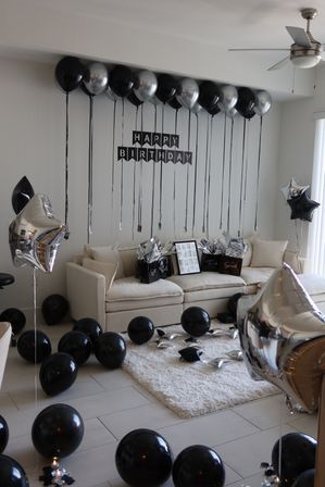 Modern living room decorated for a black-and-silver birthday: beige sofa with gift bags, a 'Happy Birthday' banner, ceiling-tethered black and metallic silver balloons and star-shaped mylar balloons scattered on the floor and rug.