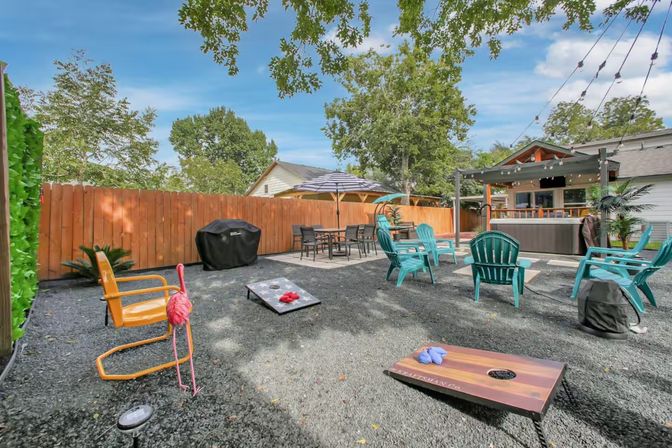 Suburban backyard patio with teal Adirondack chairs, yellow metal chair and a playful pink flamingo decoration, cornhole boards on dark gravel, dining table with umbrella, pergola-covered hot tub, string lights and wooden privacy fence under a blue sky.