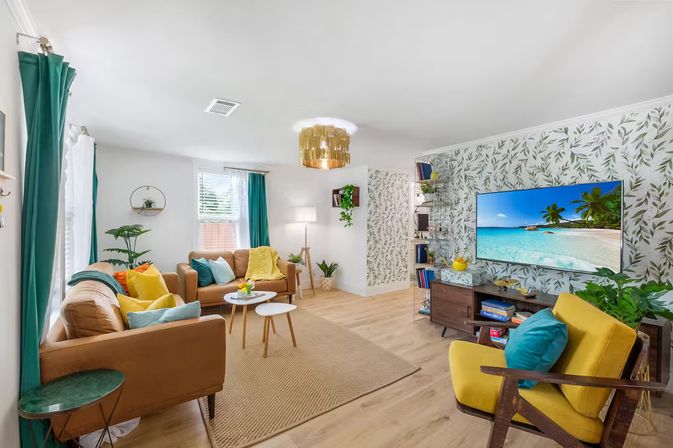 Bright modern living room with tropical leaf wallpaper, teal curtains, tan sofas, mustard accent chair, wood floors, plants and a wall-mounted TV showing a beach scene — cozy coastal-boho interior.