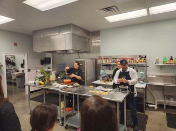 Hands-on cooking class in a commercial kitchen with instructors demonstrating fresh pasta making at stainless-steel prep tables, pasta roller, stand mixers and shelves of spices while a small audience watches.
