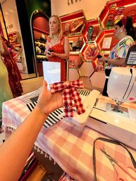 Hand holding a red gingham bow above a sewing machine on a pink-check DIY table inside a colorful accessory boutique with honeycomb shelves displaying hats, sunglasses and hair ties, while customers browse in the background.