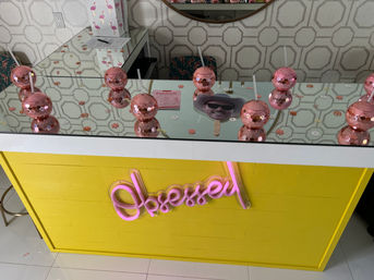 Bright yellow beauty bar counter with mirrored countertop, pink sequin double-bubble cups with straws, a handheld face fan, geometric wallpaper and neon pink cursive sign.