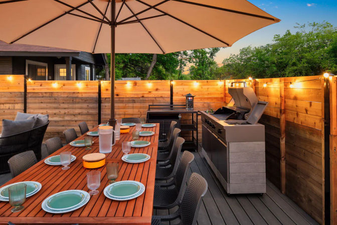 Backyard wooden deck dining area at dusk, long teak table set for eight with green plates and glassware, large patio umbrella, string lights along a wood fence and a stainless gas grill ready for al fresco cooking.