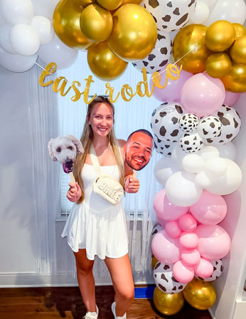 Smiling woman in a white dress at an indoor bachelorette party photo spot with a gold, pink, white and cow-print balloon arch, glitter “last rodeo” banner and dog and face cutout props