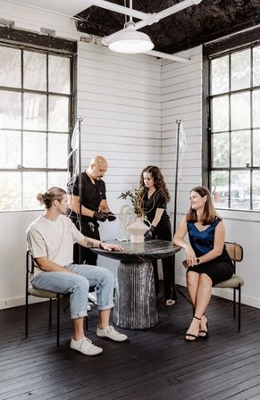 Sunlit industrial-style studio with large paned windows — two clinicians set up IV drips behind a round pedestal table while two clients sit nearby