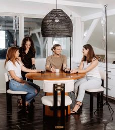 Four adults seated around a round wooden table in a bright loft-style wellness studio, chatting and sipping while two IV bags on poles hang nearby under a woven pendant light.