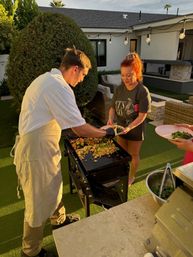 Sunlit backyard patio at golden hour with a cook serving colorful fried rice and vegetables from a flat-top griddle to guests near string lights and modern outdoor seating