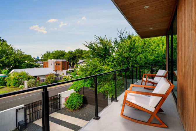 Inviting modern wood-clad balcony with glass railing and two cushioned wooden lounge chairs overlooking a leafy suburban street and contemporary homes under a bright blue sky.