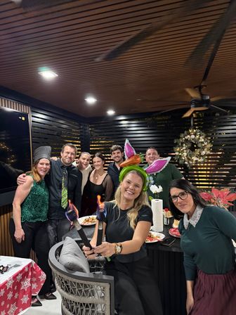 Group of friends at a festive holiday party on a covered outdoor patio—smiling adults under a wood‑slatted ceiling, woman in front holding two flaming torches and wearing a bunny‑ear balloon headband, wreath and poinsettia decorations visible.